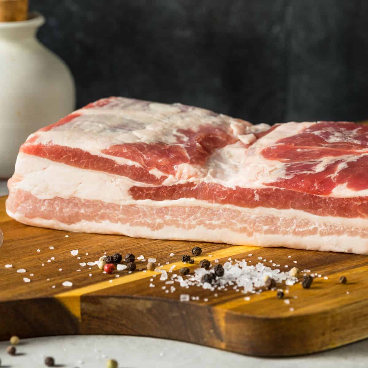 Slab of raw pork belly on a wooden cutting board with some salt and black pepper