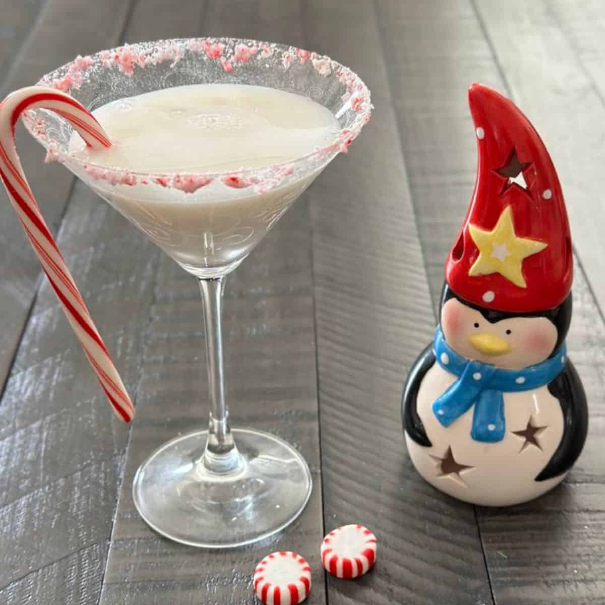Candy cane martini in a glass with a candy cane as garnish and a figurine of a penguin next to it.