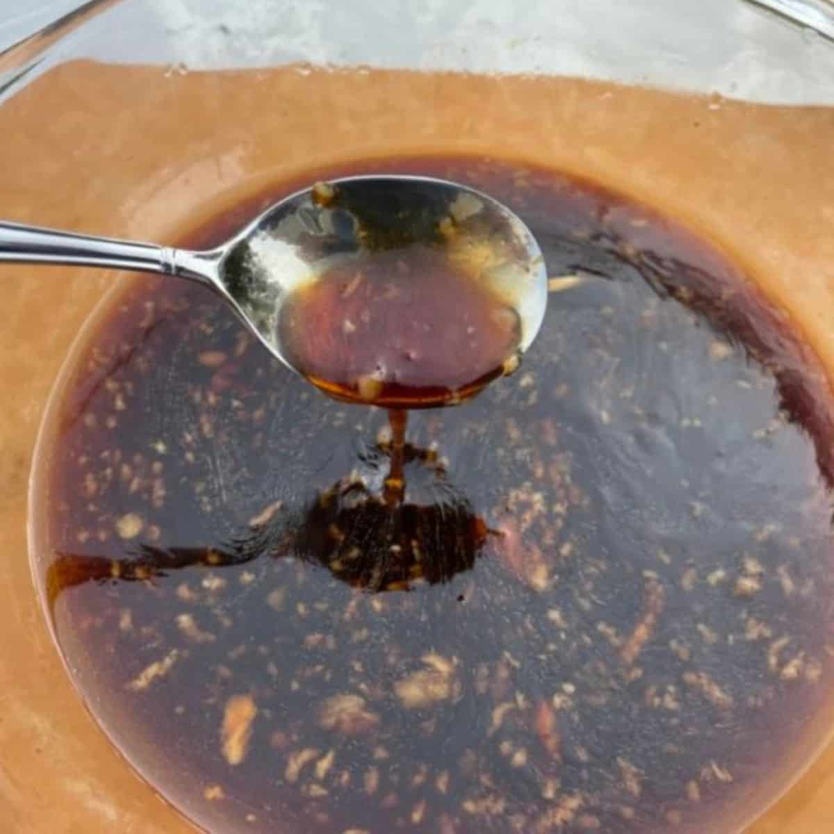 A metal spoon dipping into a glass bowl of soy honey marinade. 